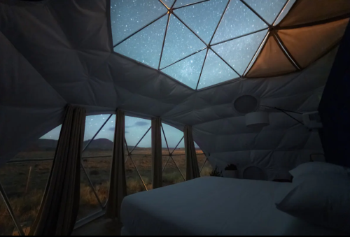 A cozy bedroom inside a geodesic dome, featuring large windows with a starry night sky view.